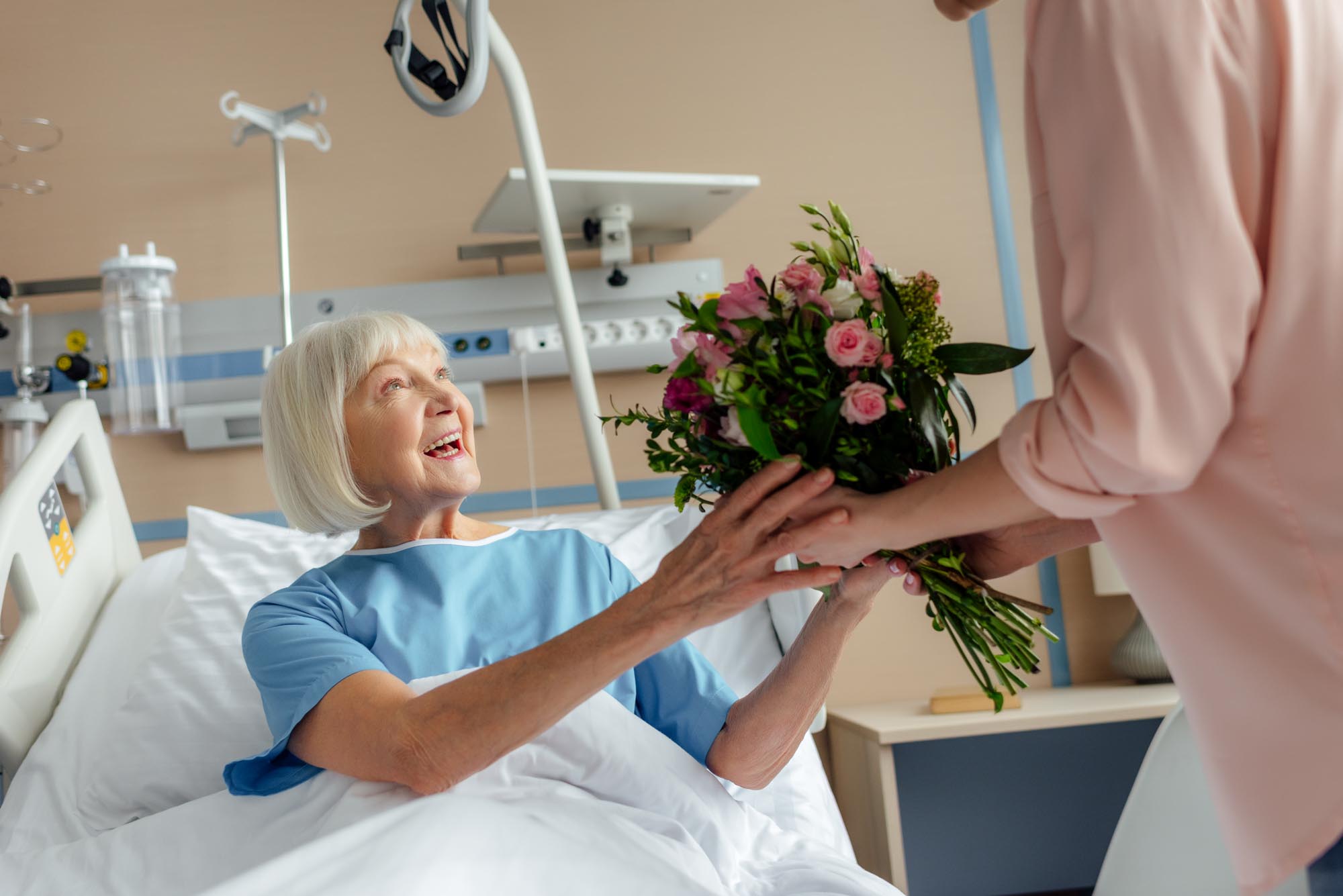 daughter presenting flowers to smiling senior woman lying in bed
